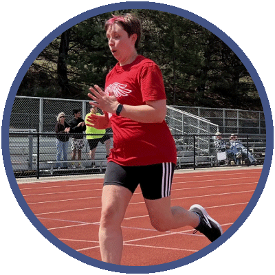 Johanna is running on a track during a race. She has short, brown hair and is wearing a bright red t-shirt and dark running shorts. She is focused on her breathing as she races.
