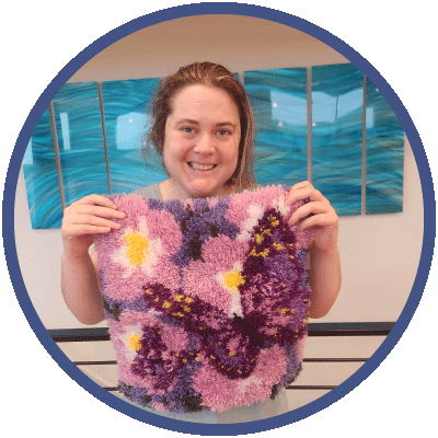 Kristin is smiling directly at the camera. She has dark brown hair and is holding up a latch hook piece she has created, with purple flowers and a butterfly on it.