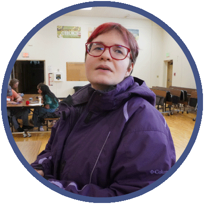 Mary is looking off into the distance as she sits at a table in a gym. She is wearing glasses and has short hair that is dyed pink. She is also wearing a dark purple winter coat.