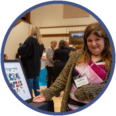 Sabrina is standing next to a painting and looking directly at the camera. She is smiling happily as she points to the framed picture. She is wearing a brown cardigan over a black and pink dress. She has long brown hair.