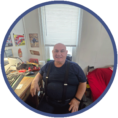 Sam is looking at the camera and smiling while sitting at his desk. He is bald and wearing a black, short-sleeved shirt.