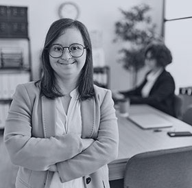 A young woman with down syndrome is standing in the foreground with her arms crossed across her body. She is standing in an office and smiling. There is an older woman sitting and working at a desk behind her.