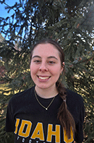 Avery is smiling directly at the camera. She is standing in front of a green leafy tree on a sunny day. She is wearing a black t-shirt with Idaho on it in yellow. She has long dark hair, in a braid, over her shoulder.