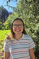 Emily is smiling directly at the camera as she stands in front of a green tree on a bright, sunny day. She has dark hair loose around her shoulders. She is wearing glasses and a striped short-sleeve shirt.