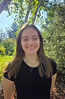 Hayley is smiling and looking directly at the camera. She is wearing a black t-shirt and is standing in front of greenery with blue skies behind her.