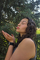 Jayda is looking off to the site with both of her hands up as if she's blowing a kiss. She is wearing a black top and standing in front of a green tree.