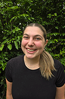 Sofia is standing in front of an evergreen tree outside. She is smiling and looking directly at the camera. She has long blond hair, pulled back into a low ponytail and is wearing a black t-shirt.