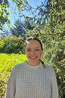 Bella is standing outside in front of a tree. She is smiling at the camera. Her dark hair is pulled back away from her face and she is wearing a light-colored sweater.
