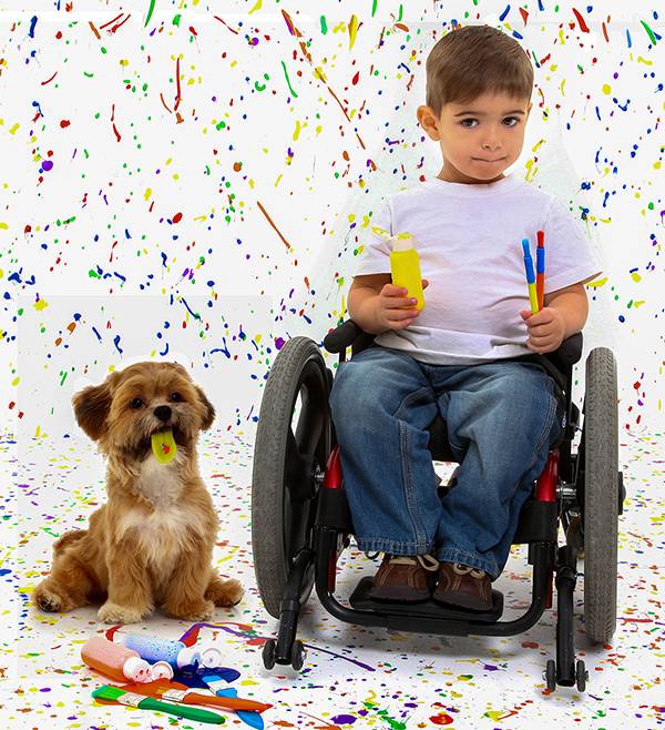 Young boy in a wheelchair with his dog. The boy is holding paint brushes and paints and sitting in front of a painted white poster.