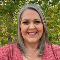 Allyson is standing in front of a white wall. She is wearing a pink top. Her silver hair is curled and pulled into a pony tail. She is smiling