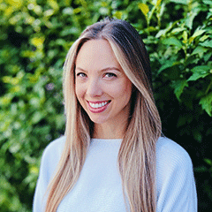 Jessi is standing in front of a leafy green bush. She is looking and smiling at the camera. She has long, blond hair falling over her shoulders and is wearing a white top.