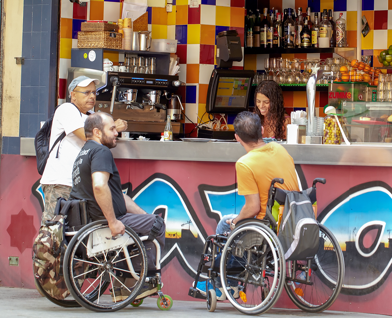 Two men in wheelchairs ordering coffees at a street stand with an onlooker standing next to them.